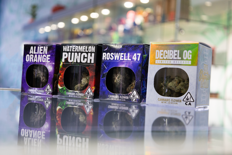 Four boxes of Decibel Gardens cannabis flower—Alien Orange, Watermelon Punch, Roswell 47, and Decibel OG—displayed on a glass counter with vibrant packaging, showcasing premium California-grown strains.
