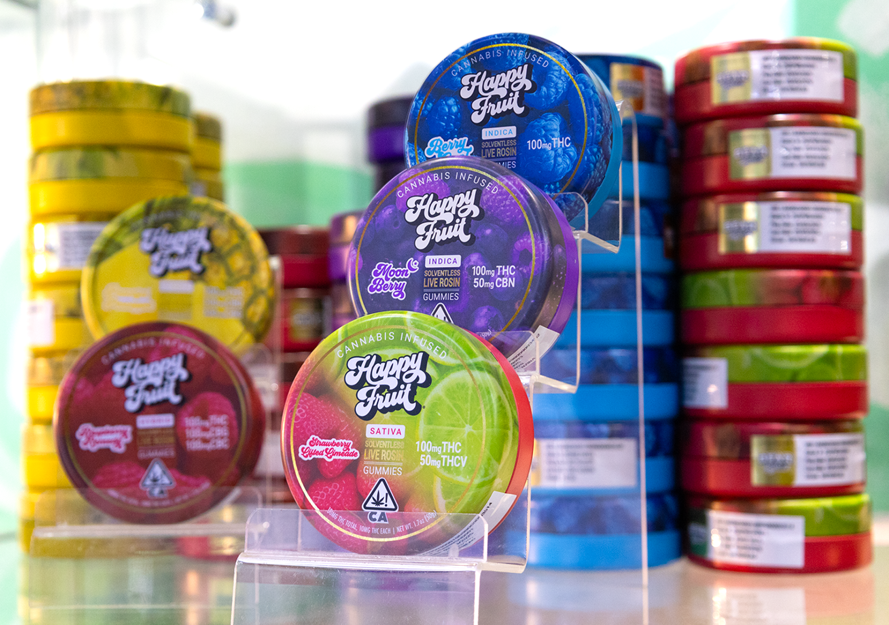 Display of Happy Fruit cannabis-infused solventless live rosin gummies in colorful round tins stacked on clear shelves. Visible flavors include Strawberry Guava (red), Moon Berry (purple), and Berry Cool (blue), with additional red, yellow, and blue tins arranged in the background.