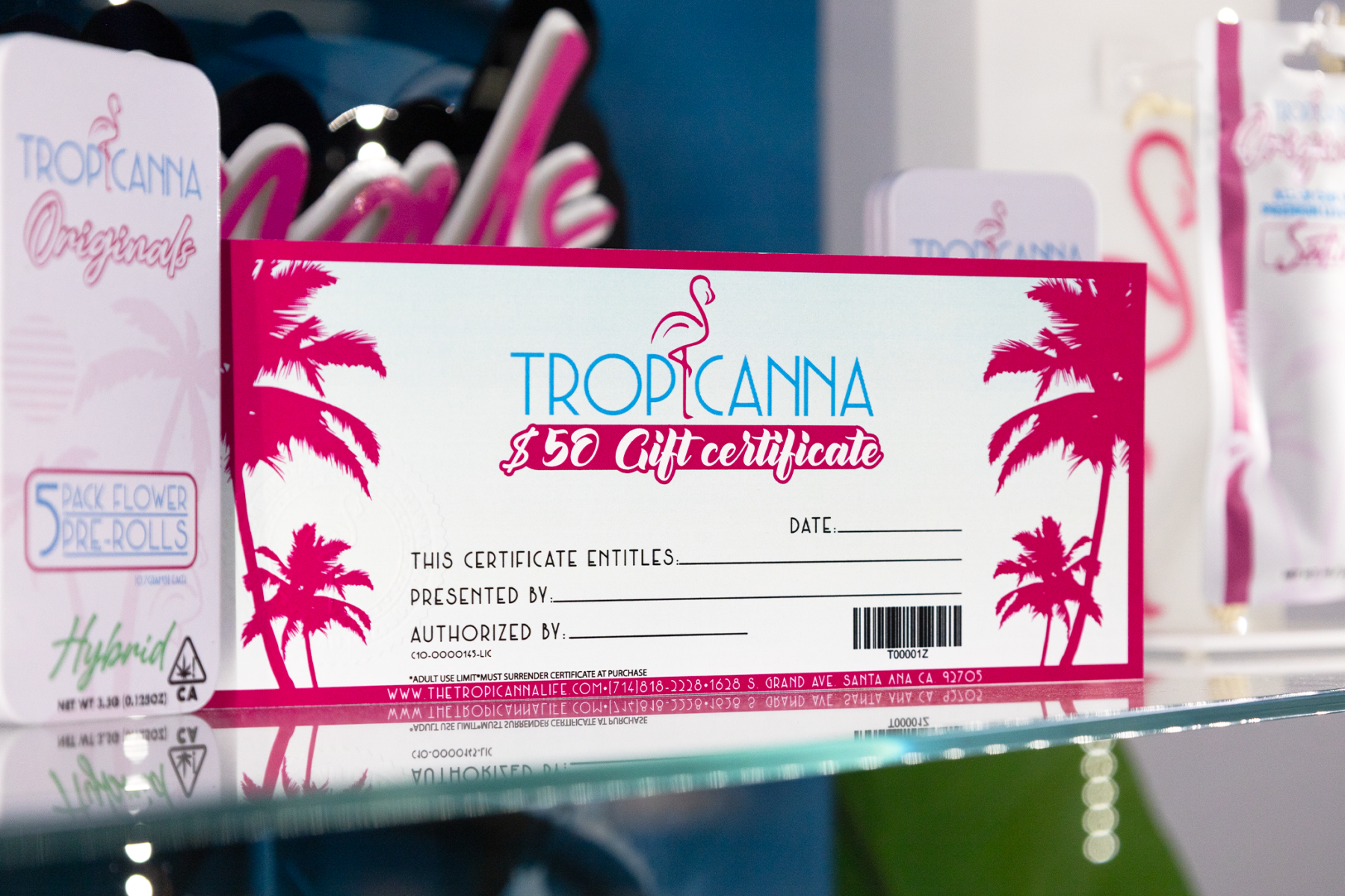 A $50 Tropicanna gift certificate displayed on a glass shelf beside Tropicanna Originals cannabis products. The card features pink palm tree graphics and the Tropicanna logo, promoting gift options available at the Santa Ana dispensary.