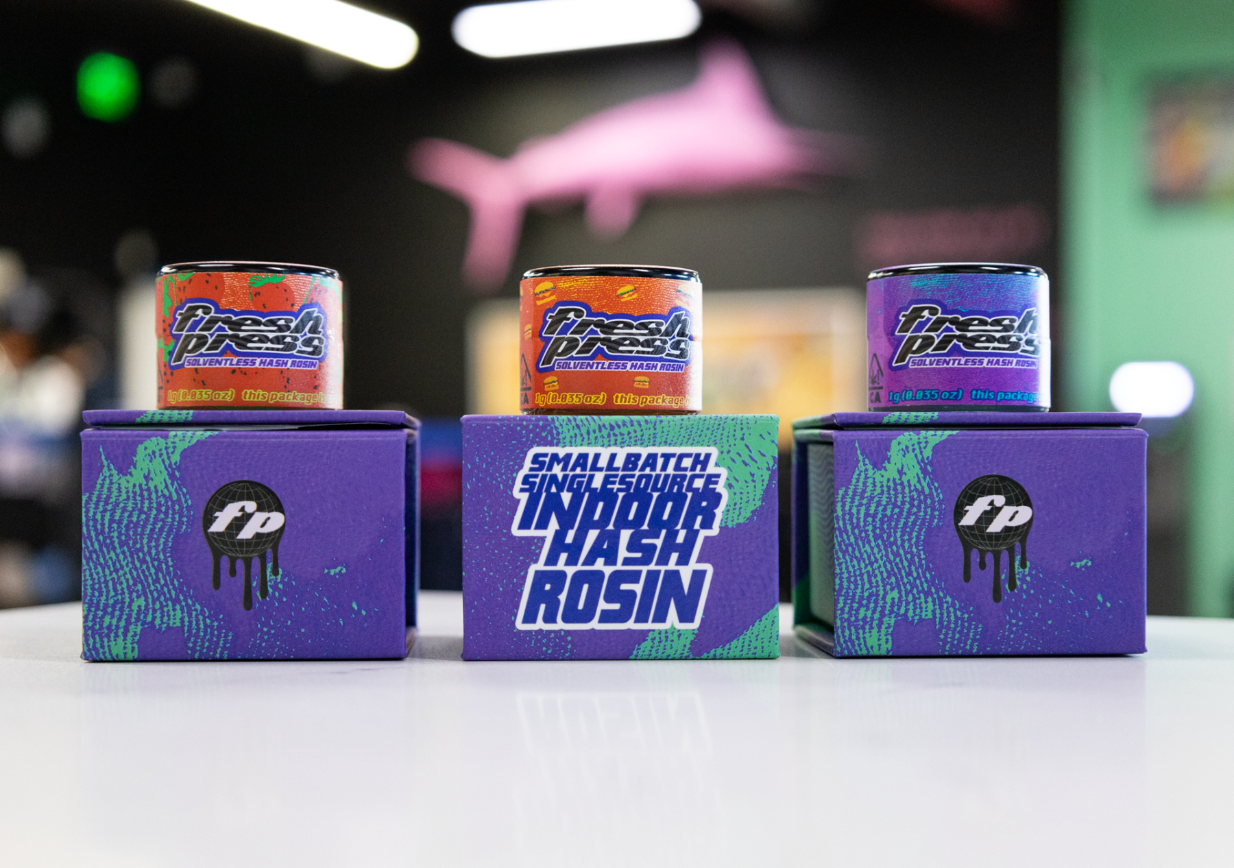 Three colorful jars of Fresh Press solventless hash rosin displayed on purple boxes labeled “Small Batch Single Source Indoor Hash Rosin,” with a blurred background featuring a pink shark wall decoration.